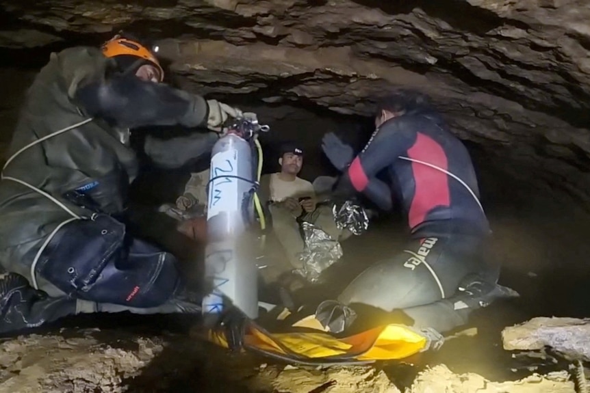Two divers squat in a section of a cave with a low ceiling, talking to a man without a shirt.