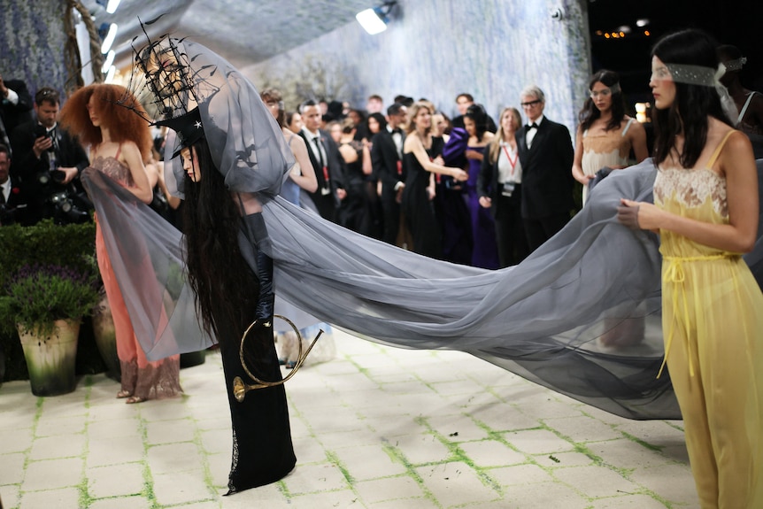 Madonna is at the Met Gala wearing a black gown with a hat shaped like a ship and a long grey train.