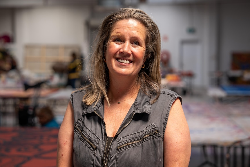 A woman with blonde hair wearing a charcoal jumpsuits stands in an art studio smiling at the camera. 