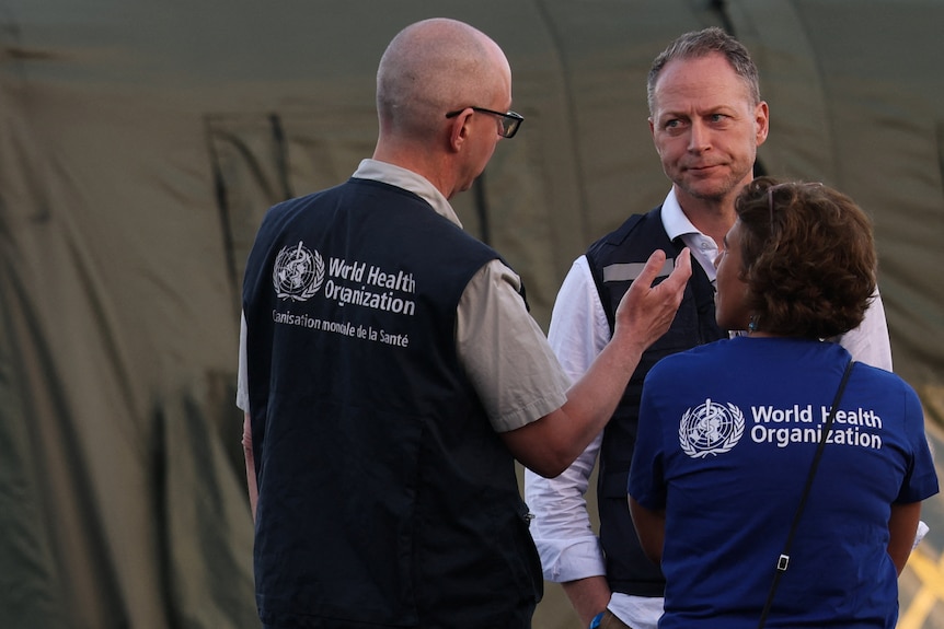 Three people talking, wearing vests and shirts with the words 'World Health Organization' on the back. 