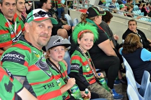 A man wearing a Rabbitohs jersey, sitting next to his grandkids in a grandstand