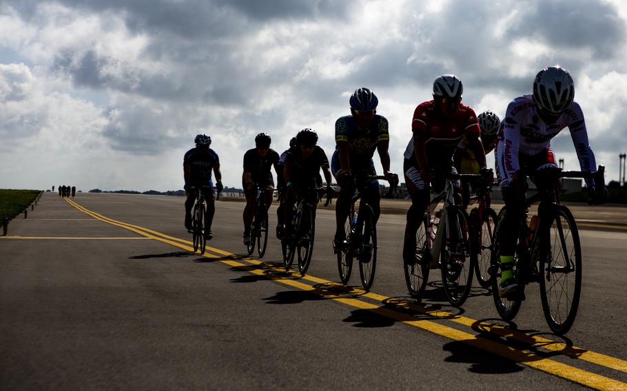 A pack of cyclists in helmets and racing kit ride down a road in silhouette under a cloudy sky.