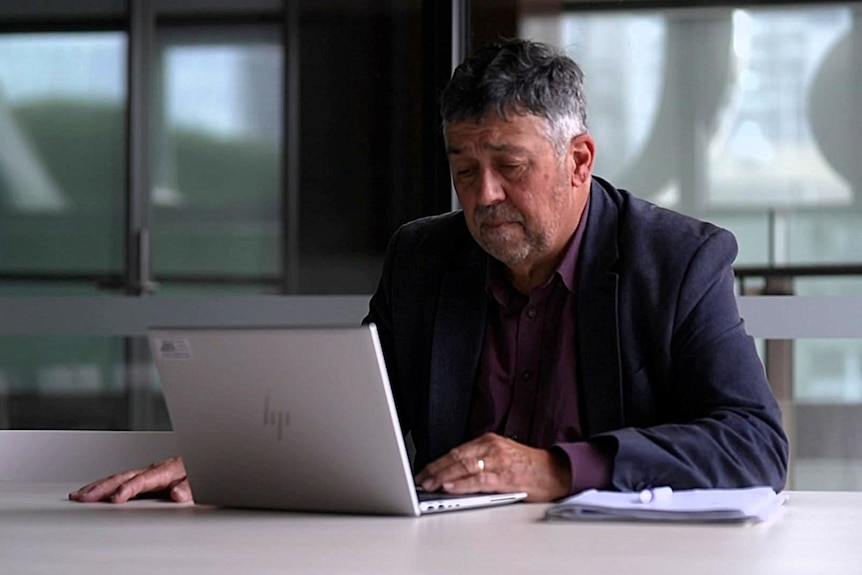 A middle aged man with short grey hair sitting at a table looking at a laptop