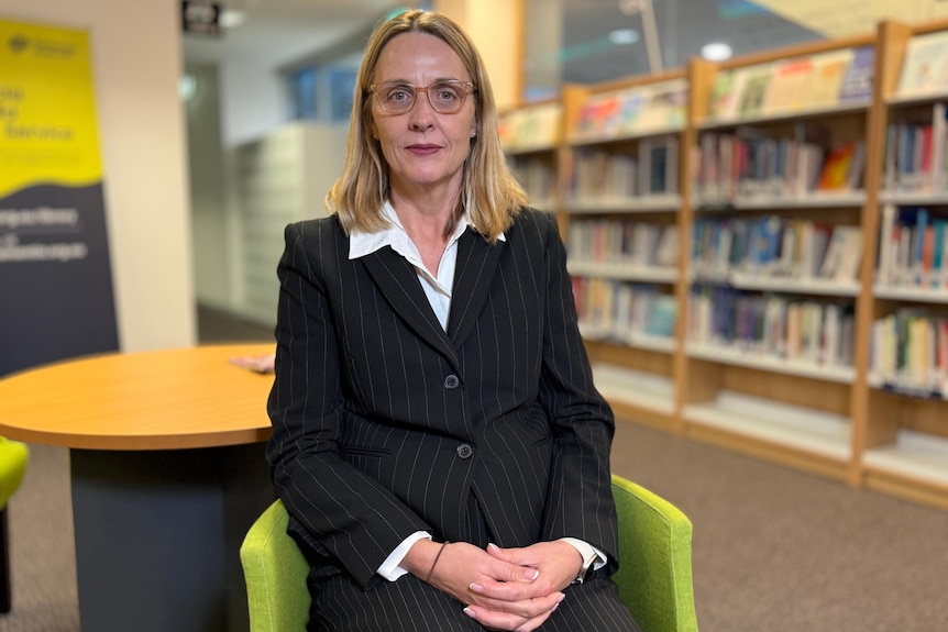 A woman in a black suit and mid-length blonde hair sits on a green chair with shelves of books behind her.