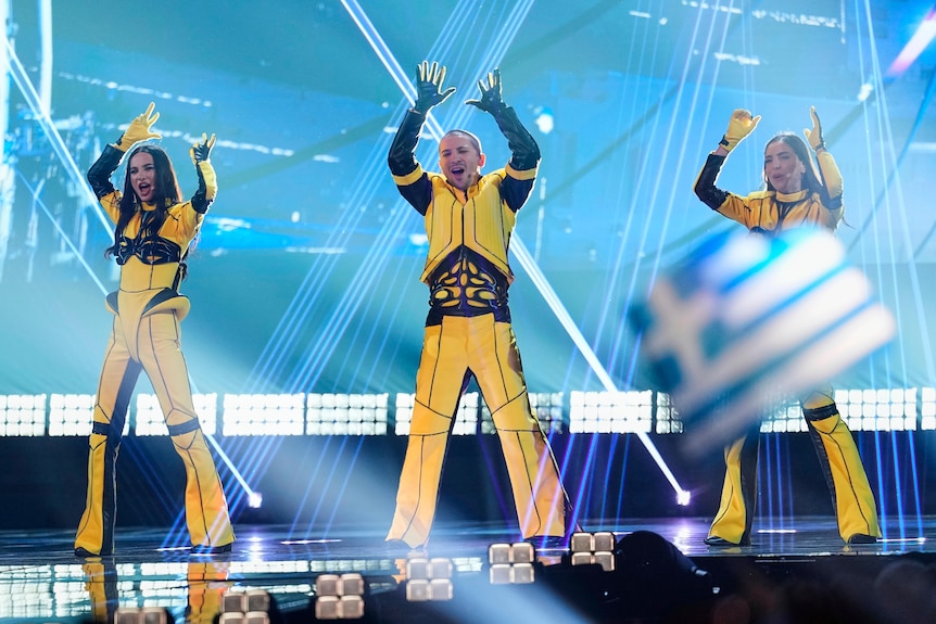 three singers in yellow and black jumpsuits clap on stage