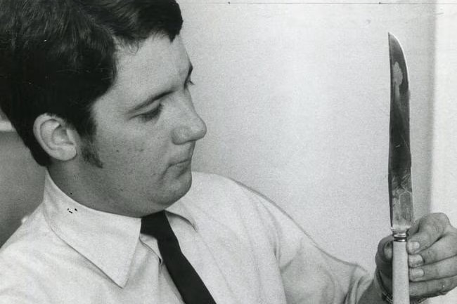Black and white photo of a man in a shirt and tie holding a large knife