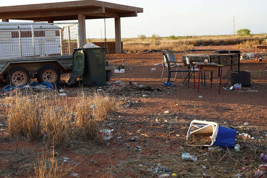 A wide shot of a caravan, trailer, and upturned esky strewn with rubbish.