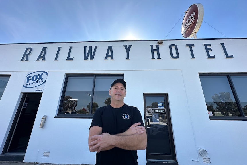 A man wearing a cap crosses his arms out the front of a hotel on a sunny day.