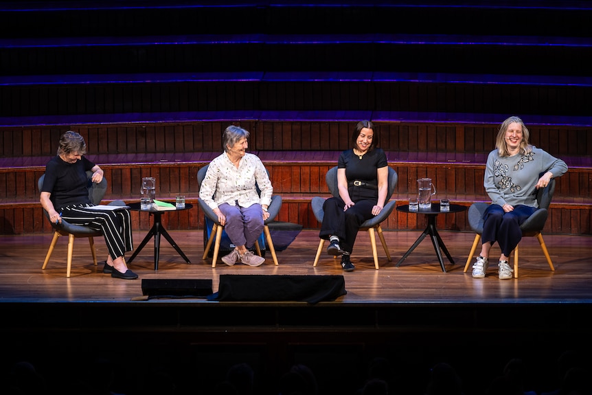 Fran Kelly, 68, Helen Garner, 83, Sarah Krasnostein, 48, and Chloe Hooper, 52, sit on stage in a row, smiling at each other.