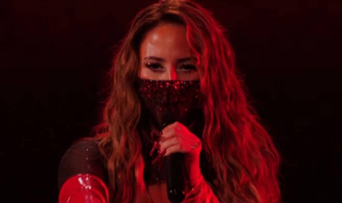 Brunette woman holding a microphone and wearing black mask.