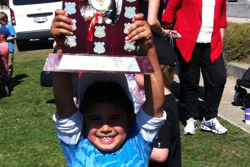 Kid holding a trophy up above his head and grinning.