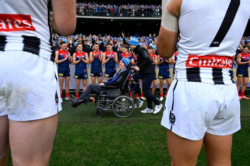 Neale Daniher is wheeled between Collingwood and Melbourne AFL players.