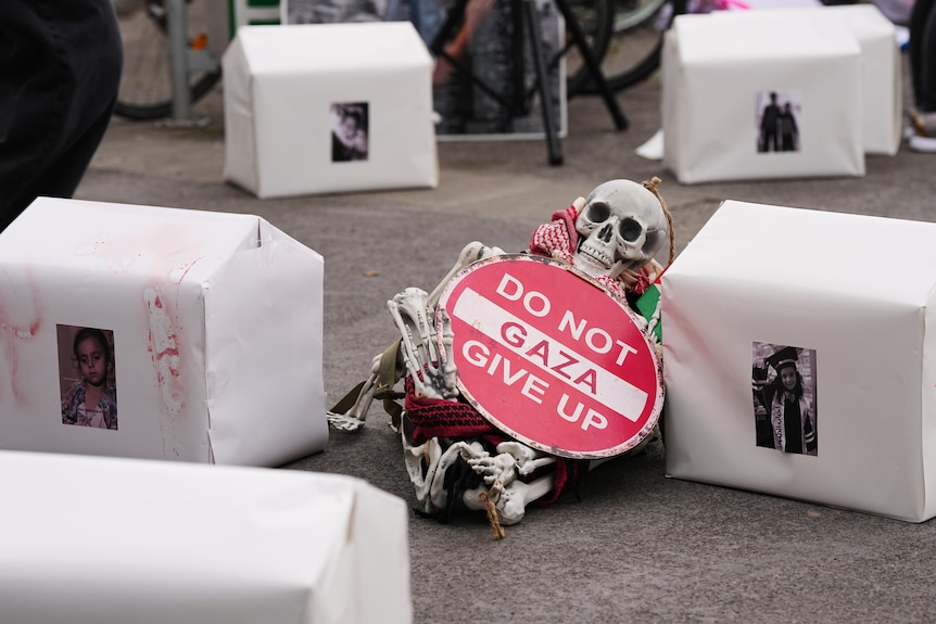 white boxes suround a fake skeleton which holds a sign 'Gaza: do not give up'