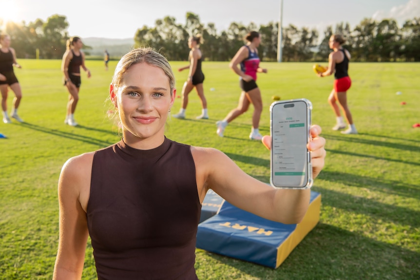 A woman holds a phone up to the camera while standing on a football field.
