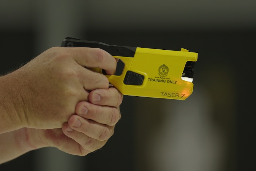 A man's hands hold a yellow taser device that is branded with "NSW Police Force TRAINING ONLY".