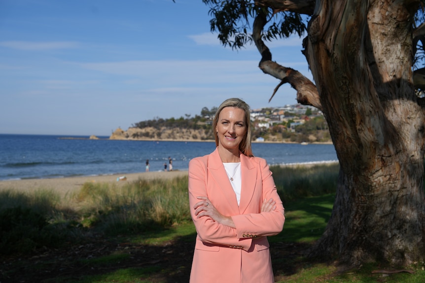 A woman wearing a white shirt and peach blazer has her arms crossed and smiles, beach in the background