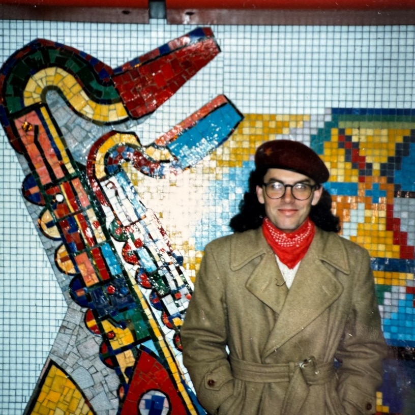 An old photo of a young man with long hair spreading behind his head, wearing glasses, in front of a colourful saxophone mural.
