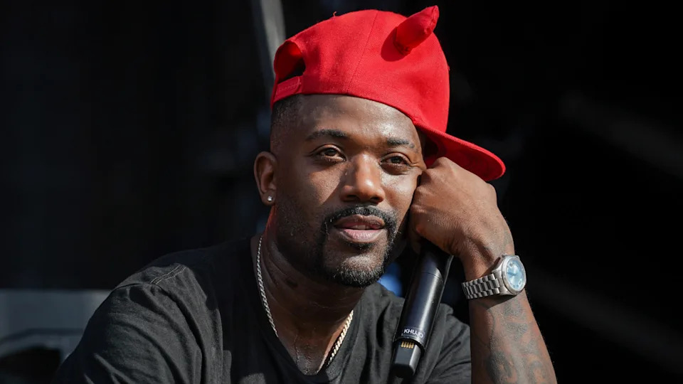 Ray J in a red cap with horns, holding a microphone, wearing a black shirt and a watch, looking thoughtful.