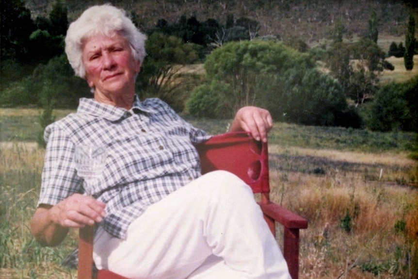 An older woman with short grey hair reclines on a red camping chair in an unidentified paddock.