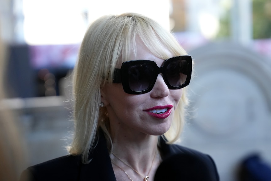 A blonde woman in a black sit with red lipstick and dark sunglasses
