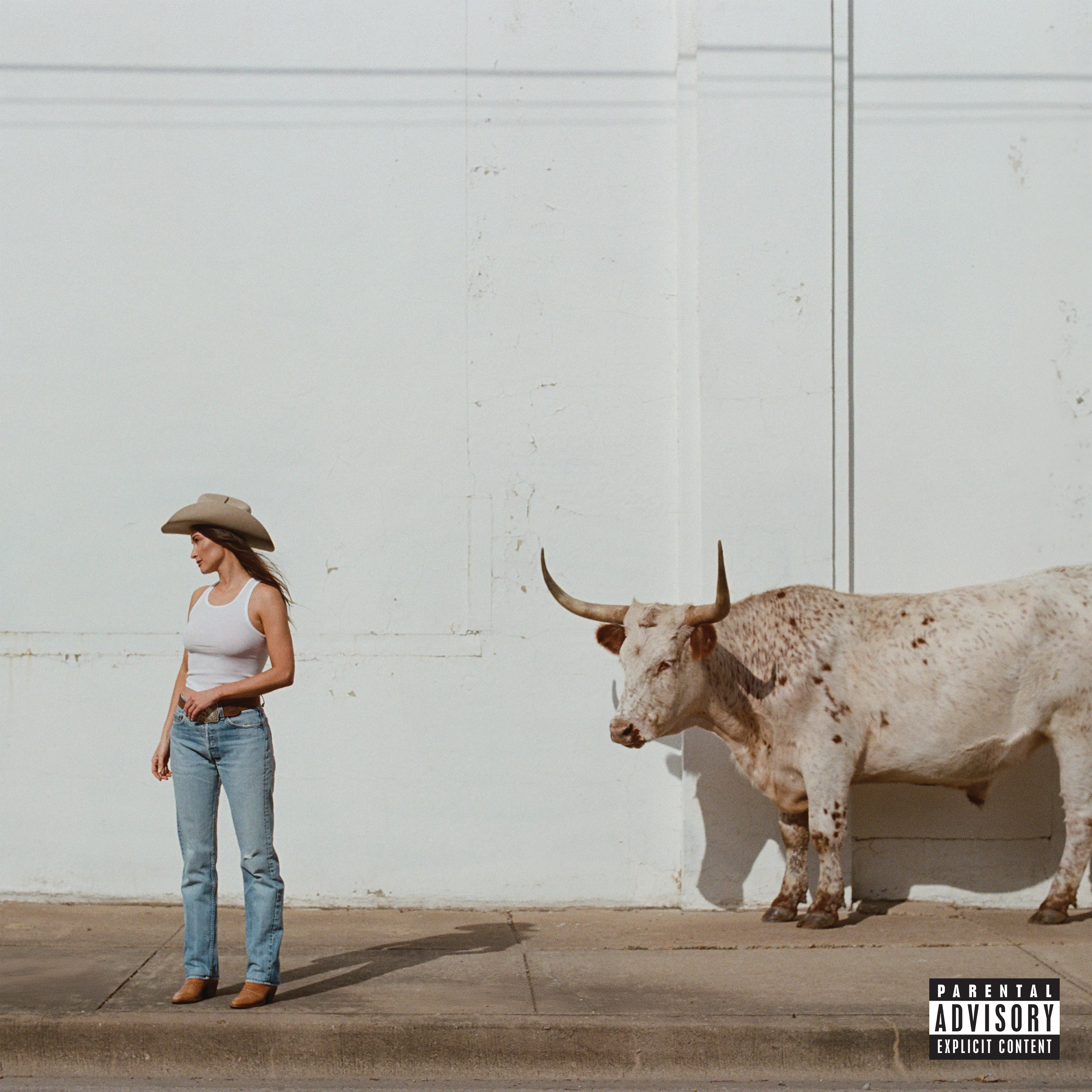 White woman in cowboy hat, blue jeans & tank top stands on street with a spotted beige bull behind her.