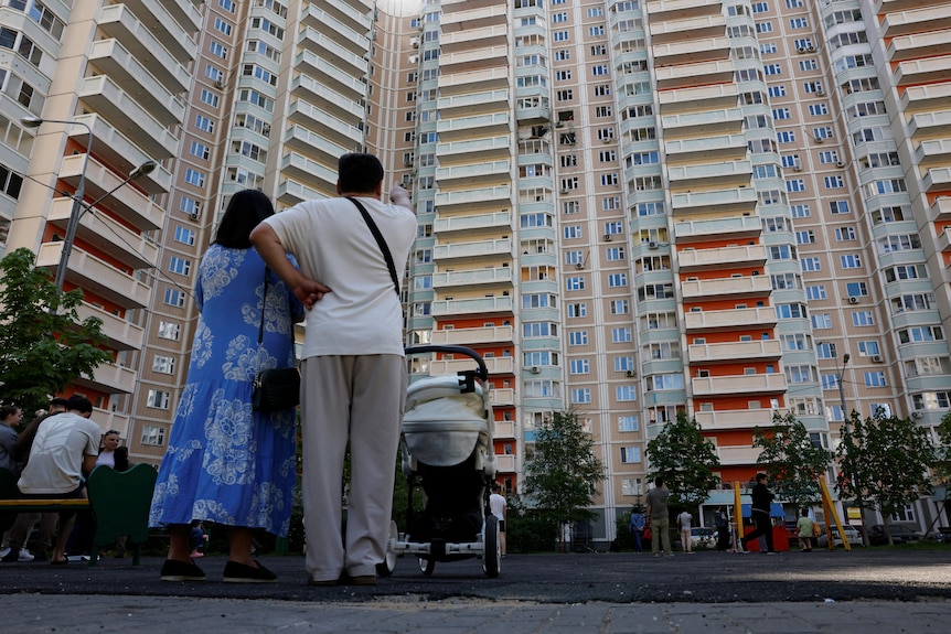 A couple stand in front of a large apartment building, pointing at a damaged apartment halfway up.