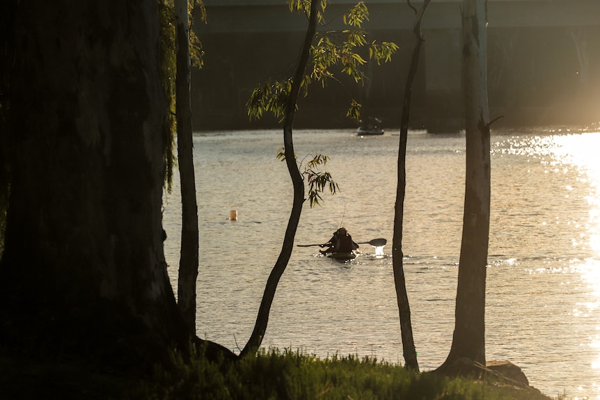 View of river looking through trees, reflection of sun bounces off the river, figures can be seen in a boat with paddles.