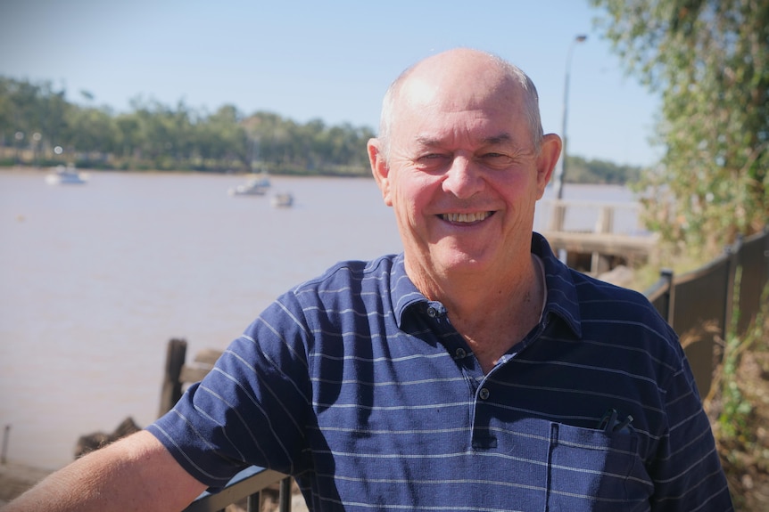 A man in a blue shirt smiles as he stands near a river.