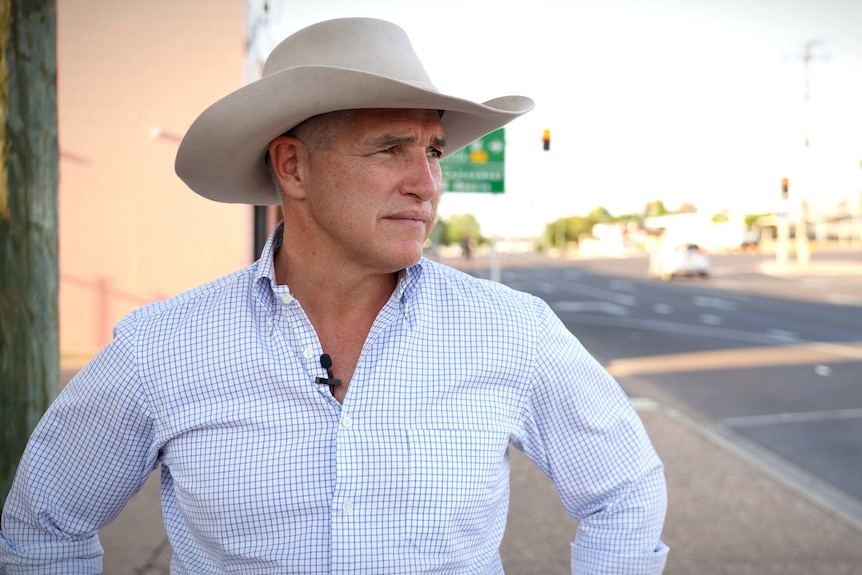 A man standing on a bitumen street wearing a button up shirt and large hat, he has a serious expression on his face.