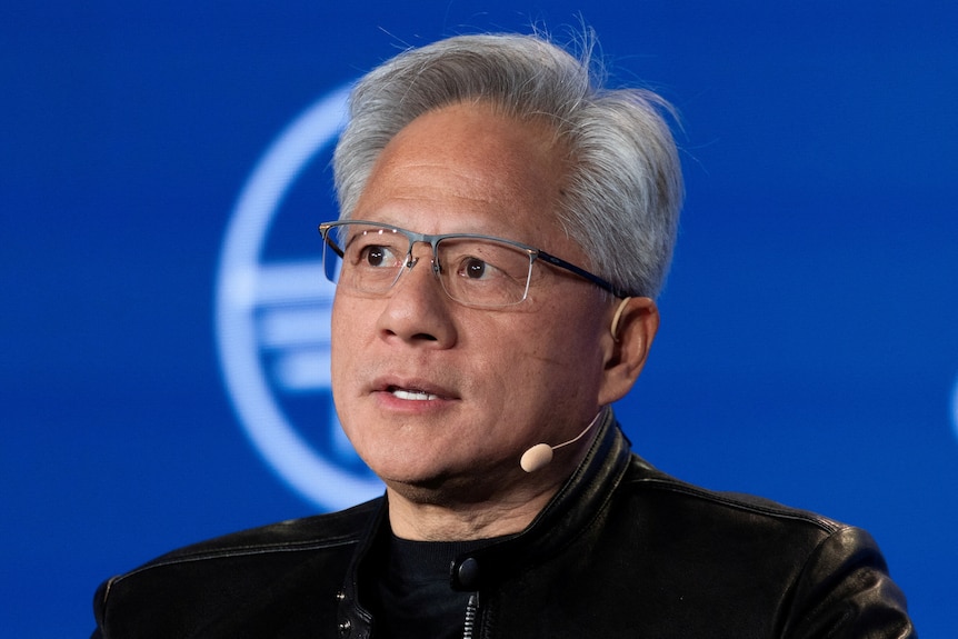 Huang looks off with a thoughtful expression as he speaks on a stage.