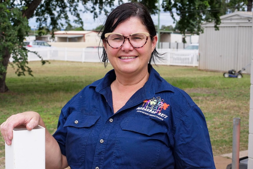 A smiling, dark-haired woman in glasses and a branded button-up shirt stands in outdoors.