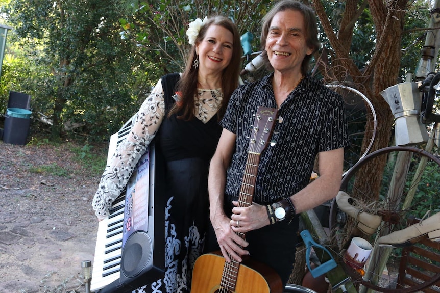 Ursula wears a black dress, holding a keyboard under her arm, while Mick, in a blue shirt, stands with an acoustic guitar.