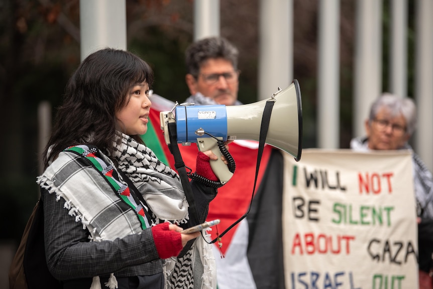 Woman wearing a Palestinian scarf speaks into a megaphone in front of other protesters outside court.