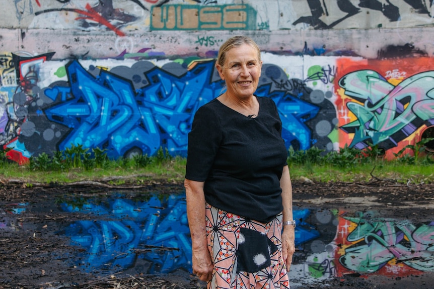 A woman in a black top smiles while standing in front of a wall covered in graffiti.