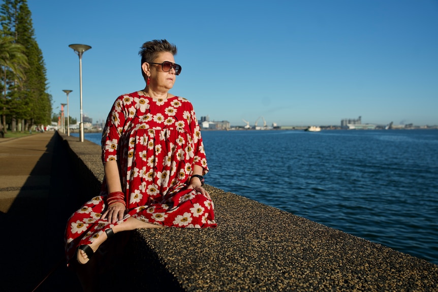 A woman sits by the water wearing a red dress with floral print.