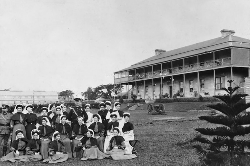 A black and white image of a group of nurses sitting on a lawn posing for a photo in front of a large two-story building.