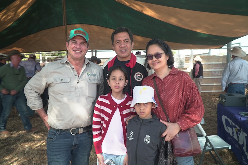 White man standing next to an Indonesian man and woman, young girl and boy, standing on hay ground, casual clothes.