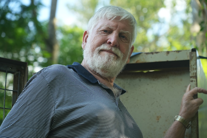 A man with white hair and beard looking down at camera. He is leaning against a rusted metal cabinet in his backyard.