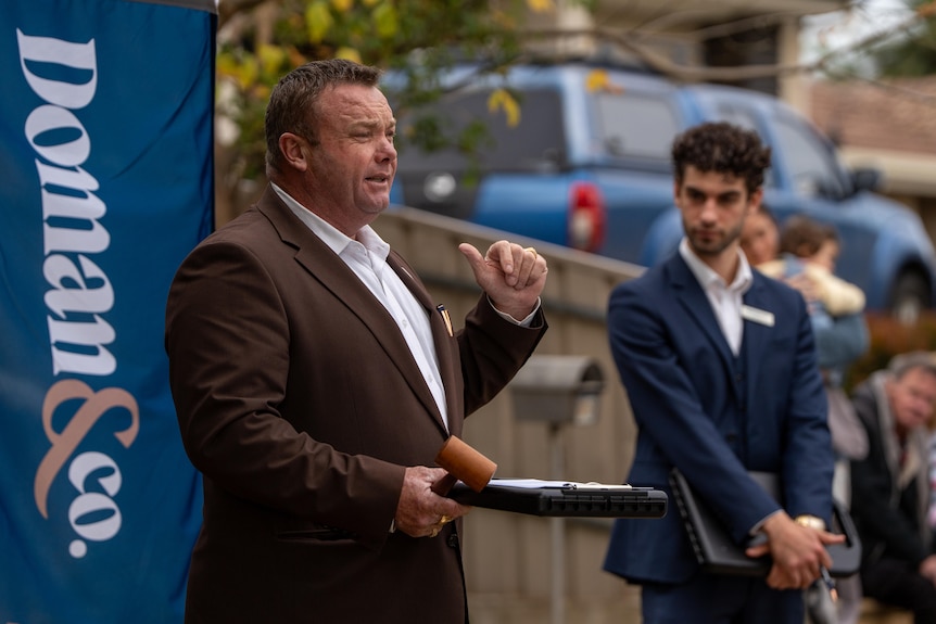 A man wearing a brown suit gesticulates at a banner