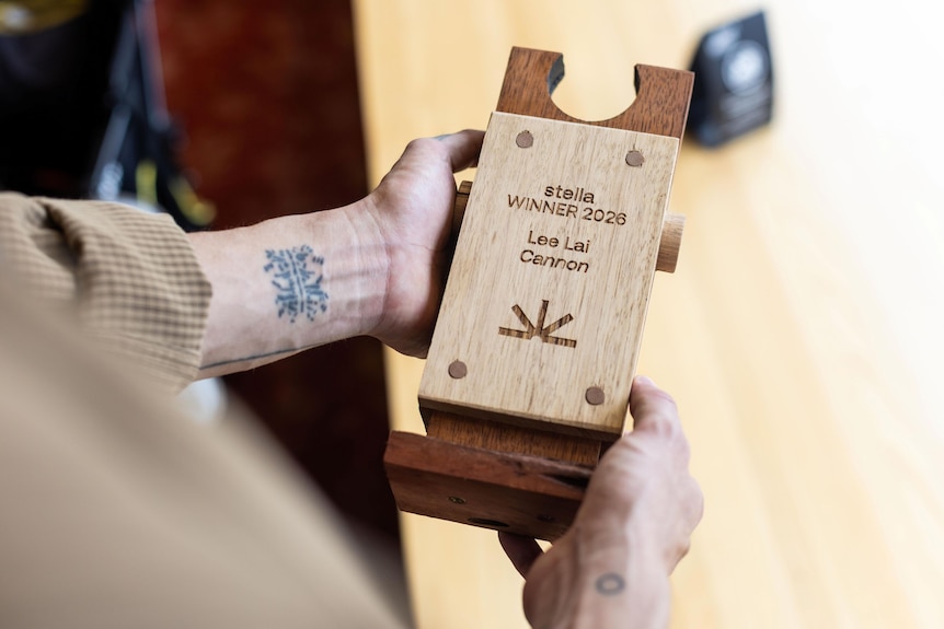 A person's hands holds a wooden trophy inscribed with the words: 'Stella winner 2026, Lee Lai, Cannon'.