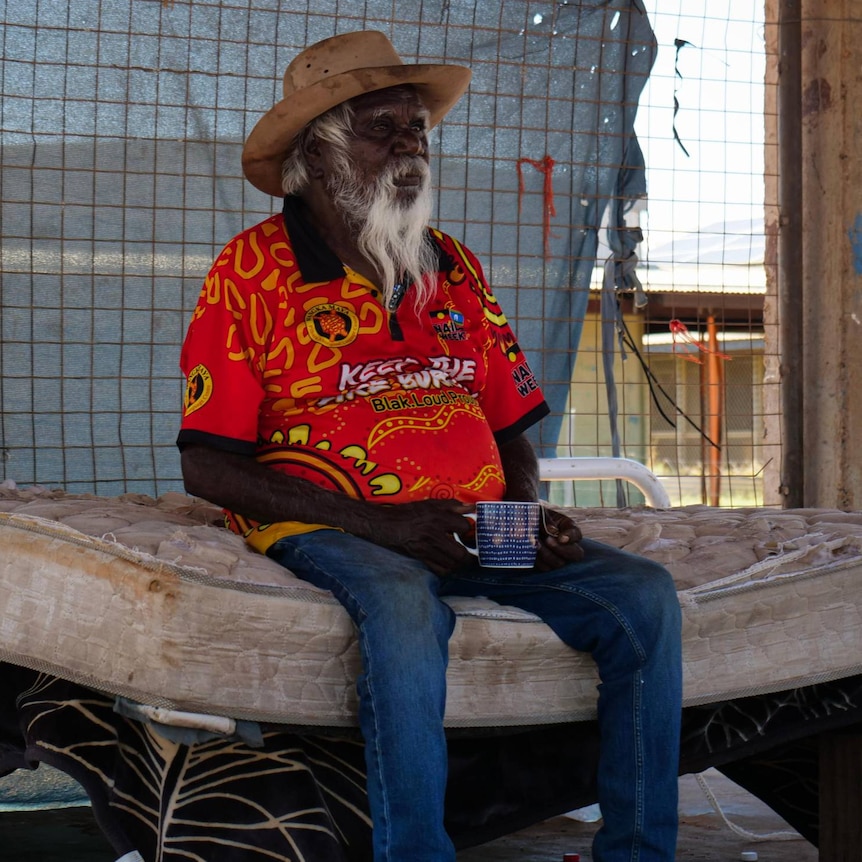 A wide shot of an elderly Aboriginal man with a white beard sitting on a bare cot.