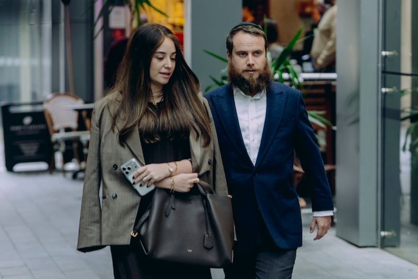 Ms Gutnick with long brown hair walks next to a man wearing a kippa and a suit past a cafe.