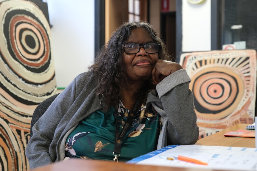 Audrey Brumby, surrounded by Aboriginal paintings, looks at the camera.