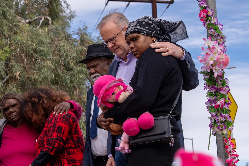 White man in suit standing with two Aboriginal people, mand and woman, among pink flowers along a fence line.