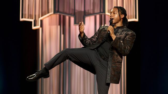 Josh Johnson doing a karate kick on stage with a chandelier installation behind him in 'Josh Johnson: Symphony' 