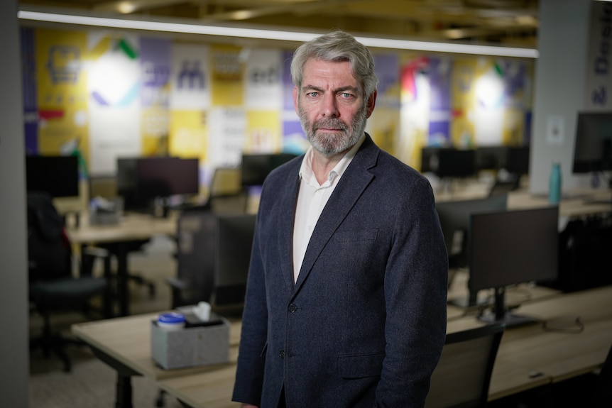Peter is serious, has grey hair and beard and moustache, wear blue suit, white shirt, background blurred.