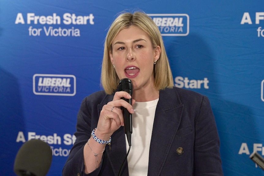 A woman with chin length blonde hair in a dark jacket and white shirt holds a microphone in front of a Liberal Victoria sign.