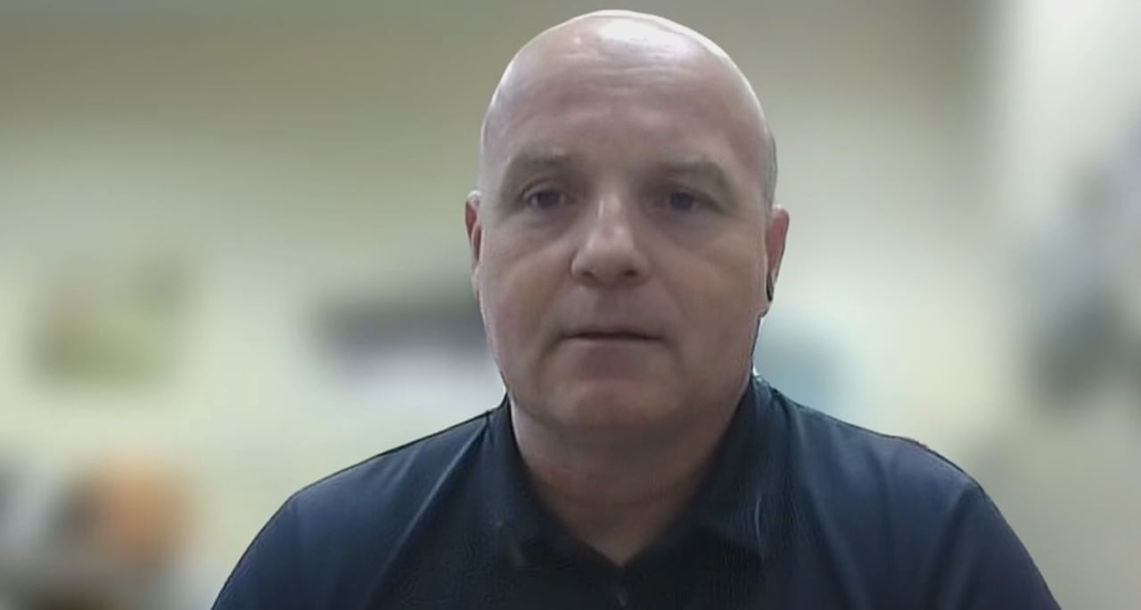 A man in a dark shirt sits and speaks in front of a blurred-out background.