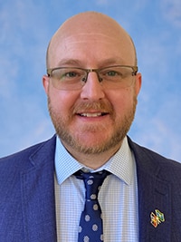 A smiling, dbearded man in a dark blazer and tie.