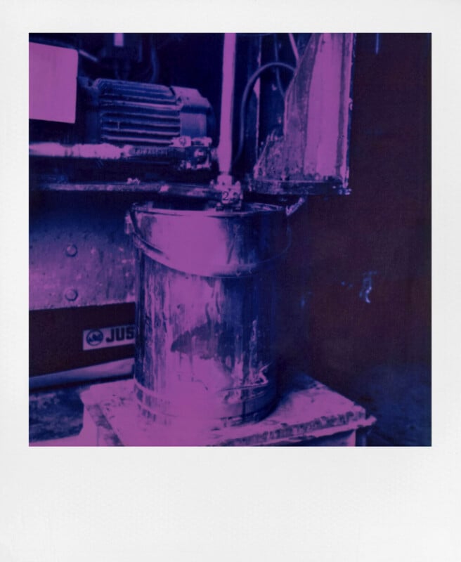 A metal bucket sits below a large industrial machine, illuminated by purple and pink lighting. The scene appears to be part of a workshop or factory setting, with visible grime and mechanical components.